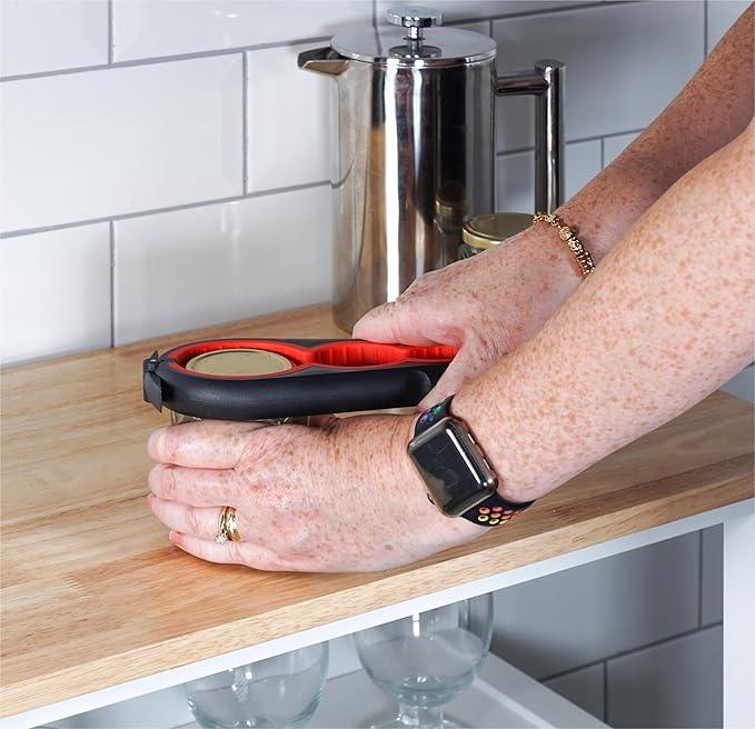 Hands using a jar opener tool on a kitchen counter to open a jar lid