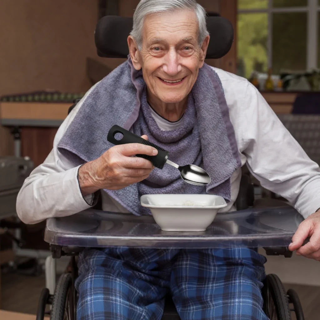 Elderly man in wheelchair using adaptive easy-grip spoon to eat from bowl at table