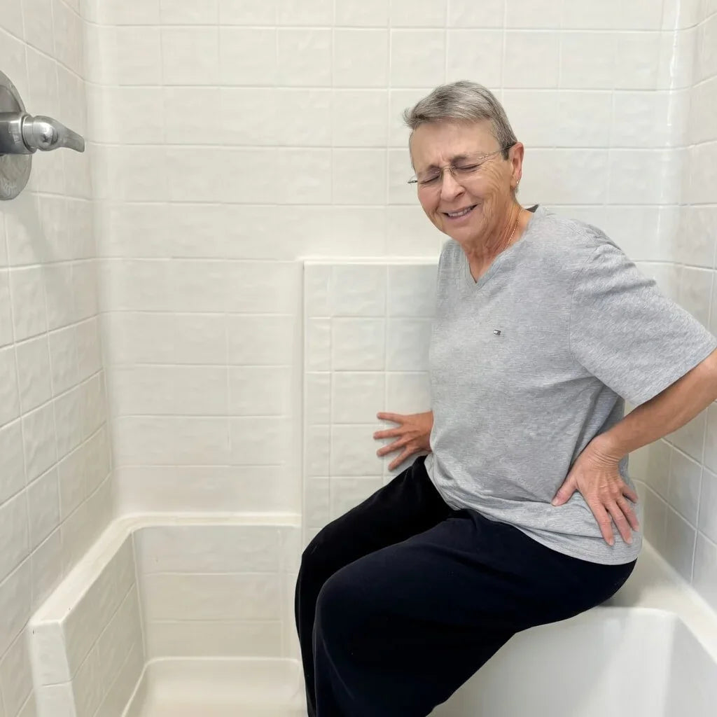 Older person in gray shirt sitting in shower with hand on lower back, showing discomfort