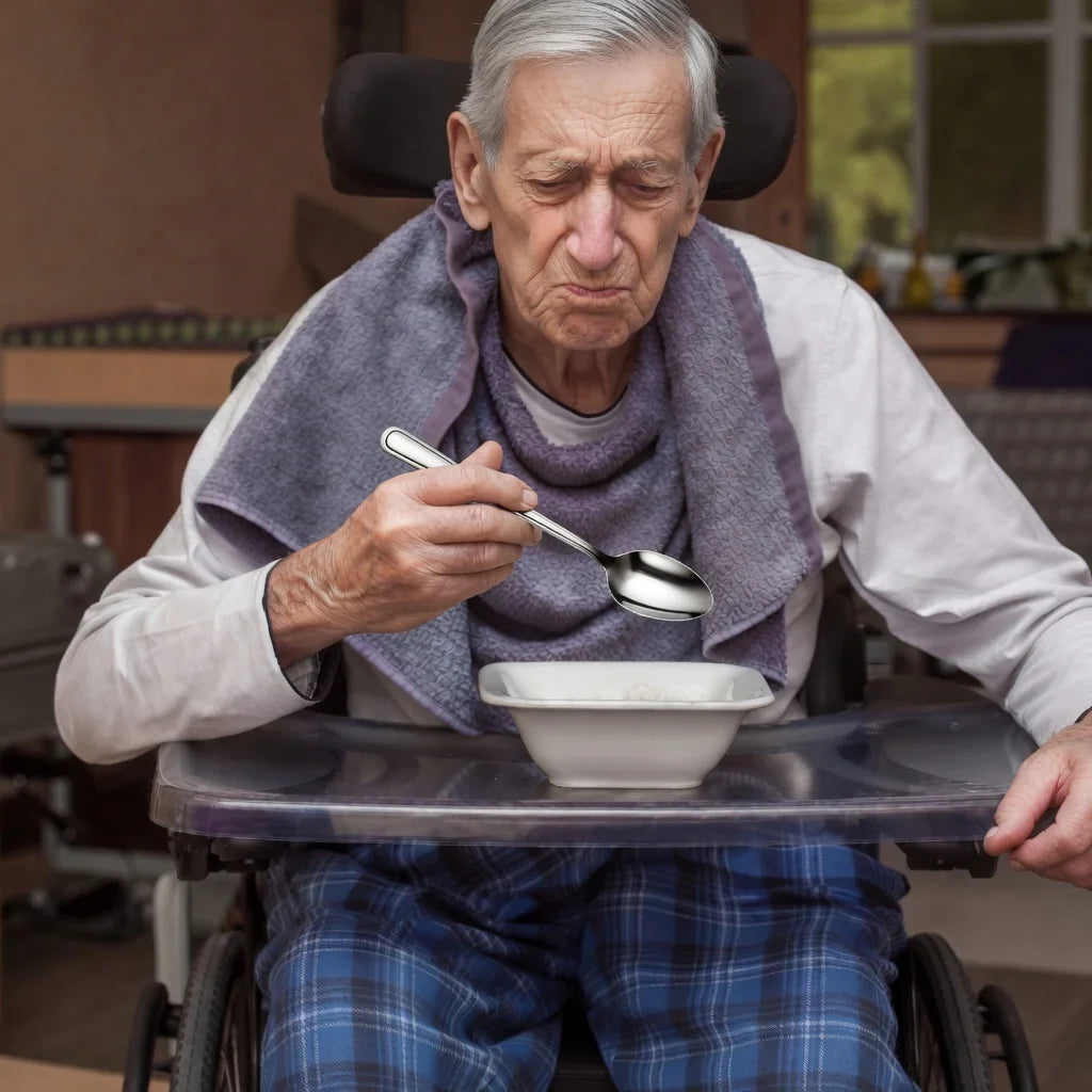 Elderly man in wheelchair eating with adaptive spoon at table, wearing a bib